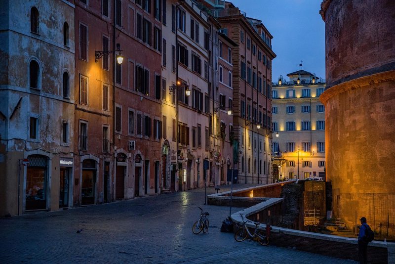 Illustration: A narrow street in Rome at dusk with glowing streetlights